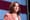 Kamala Harris in a pink jacket and pearls, with an American flag behind her